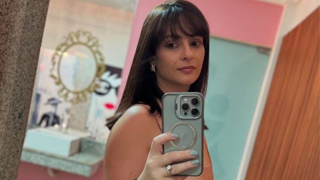 Mulher de cabelos castanhos, com franja, tira uma selfie em frente ao espelho usando um celular; ela veste roupa sem mangas e está em um ambiente interno com paredes em tons de rosa e azul e objetos decorativos ao fundo.