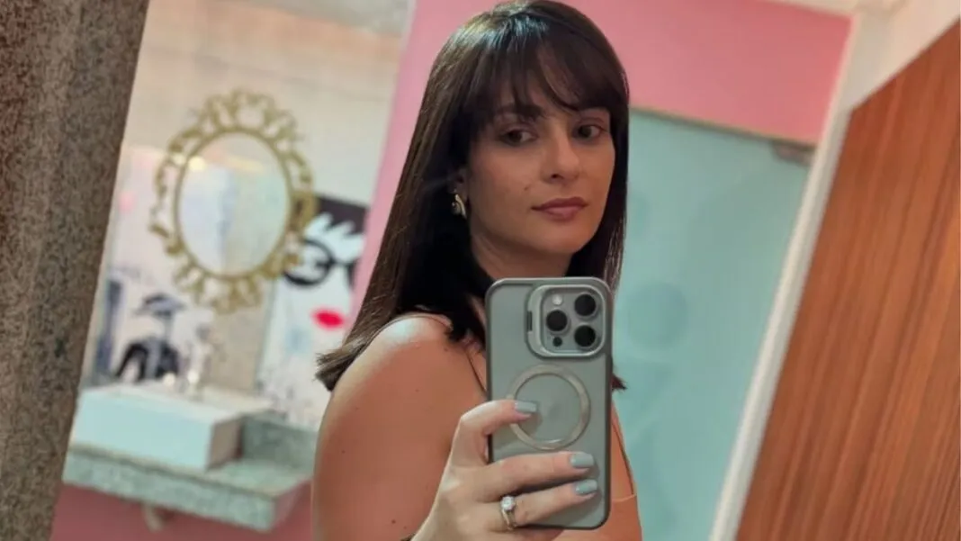 Mulher de cabelos castanhos, com franja, tira uma selfie em frente ao espelho usando um celular; ela veste roupa sem mangas e está em um ambiente interno com paredes em tons de rosa e azul e objetos decorativos ao fundo.