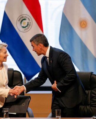 Autoridades participam de um encontro oficial: uma mulher de blazer claro cumprimenta um homem de terno escuro com um aperto de mãos, enquanto outros líderes observam sentados à mesa; ao fundo, bandeiras de países da América do Sul e do Brasil indicam um contexto diplomático ou internacional.