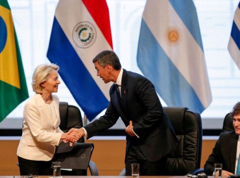 Autoridades participam de um encontro oficial: uma mulher de blazer claro cumprimenta um homem de terno escuro com um aperto de mãos, enquanto outros líderes observam sentados à mesa; ao fundo, bandeiras de países da América do Sul e do Brasil indicam um contexto diplomático ou internacional.