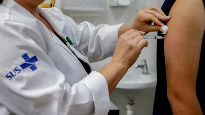 Enfermeira vestida com as roupas do SUS aplica uma vacina no braço de uma pessoa.