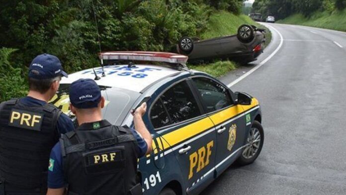 Em primeiro plano, dois policiais rodoviários federais de costas estão atrás de uma viatura da PRF estacionada na margem da rodovia. Eles observam um carro de passeio capotado às em curva sinuosa.