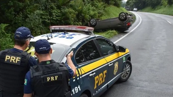 Em primeiro plano, dois policiais rodoviários federais de costas estão atrás de uma viatura da PRF estacionada na margem da rodovia. Eles observam um carro de passeio capotado às em curva sinuosa.