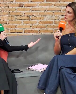 Duas mulheres estão sentadas em um sofá cinza, conversando e segurando microfones com a marca TMC — um microfone verde (à esquerda) e um laranja (à direita).