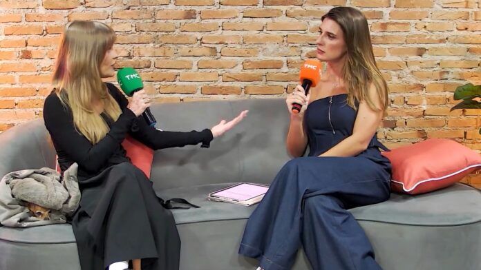 Duas mulheres estão sentadas em um sofá cinza, conversando e segurando microfones com a marca TMC — um microfone verde (à esquerda) e um laranja (à direita).