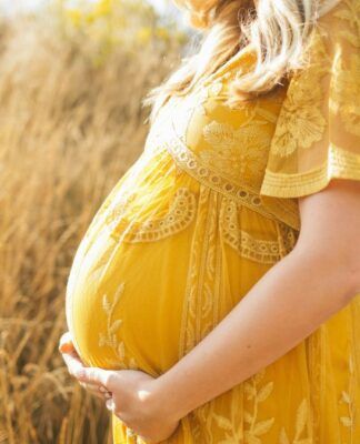 Close-up lateral do ventre de uma pessoa grávida em um campo de vegetação seca e dourada. Ela veste um vestido amarelo com detalhes em renda e bordados florais, mantendo as mãos apoiadas suavemente na parte inferior da barriga. A iluminação é solar e suave.