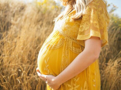 Close-up lateral do ventre de uma pessoa grávida em um campo de vegetação seca e dourada. Ela veste um vestido amarelo com detalhes em renda e bordados florais, mantendo as mãos apoiadas suavemente na parte inferior da barriga. A iluminação é solar e suave.