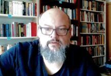 Um homem de pele clara, calvo e com barba grisalha cheia olha para a câmera. Ele usa óculos de armação escura e uma camiseta preta. Ao fundo, há prateleiras repletas de livros organizados, sugerindo um escritório ou biblioteca doméstica. A iluminação é frontal.