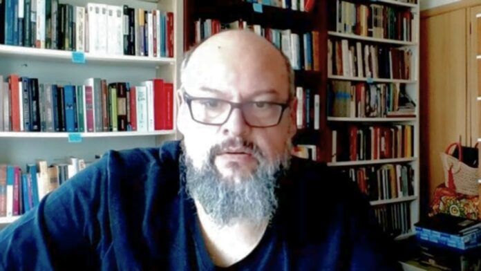 Um homem de pele clara, calvo e com barba grisalha cheia olha para a câmera. Ele usa óculos de armação escura e uma camiseta preta. Ao fundo, há prateleiras repletas de livros organizados, sugerindo um escritório ou biblioteca doméstica. A iluminação é frontal.