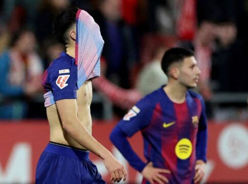 Dois jogadores de futebol em campo com uniformes azul e grená. O jogador em primeiro plano está de perfil, com a camisa puxada cobrindo o rosto, revelando o tronco. Ao fundo, outro jogador observa com as mãos na cintura. O clima sugere desânimo ou frustração.