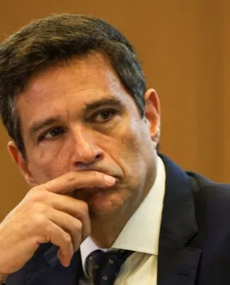 Um homem de pele clara e cabelos curtos castanhos olha fixamente para o lado com uma expressão pensativa. Ele está com o dedo indicador encostado nos lábios. Veste um terno azul-marinho, camisa branca e gravata escura com detalhes sutis. O fundo é composto por uma parede de madeira em tom mel, levemente desfocada.