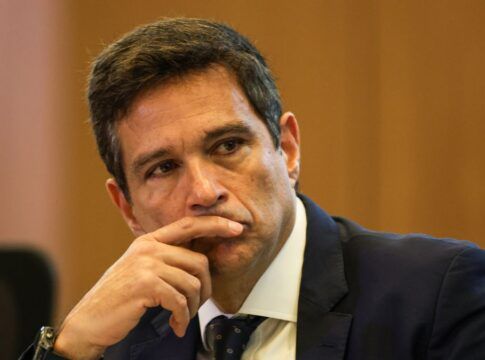 Um homem de pele clara e cabelos curtos castanhos olha fixamente para o lado com uma expressão pensativa. Ele está com o dedo indicador encostado nos lábios. Veste um terno azul-marinho, camisa branca e gravata escura com detalhes sutis. O fundo é composto por uma parede de madeira em tom mel, levemente desfocada.