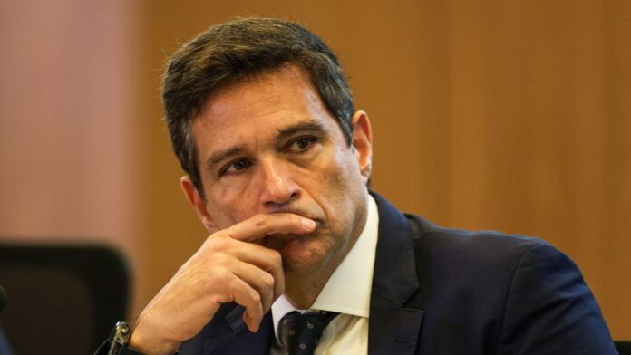 Um homem de pele clara e cabelos curtos castanhos olha fixamente para o lado com uma expressão pensativa. Ele está com o dedo indicador encostado nos lábios. Veste um terno azul-marinho, camisa branca e gravata escura com detalhes sutis. O fundo é composto por uma parede de madeira em tom mel, levemente desfocada.