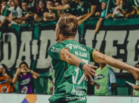 O jogador de futebol Walter Clar, da Chapecoense, é visto de costas celebrando um gol em direção à torcida. Ele veste o uniforme verde completo do clube, com os braços abertos em um gesto de triunfo. Ao fundo, a arquibancada está repleta de torcedores vibrando, com faixas e bandeiras verdes e brancas.