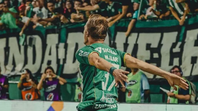 O jogador de futebol Walter Clar, da Chapecoense, é visto de costas celebrando um gol em direção à torcida. Ele veste o uniforme verde completo do clube, com os braços abertos em um gesto de triunfo. Ao fundo, a arquibancada está repleta de torcedores vibrando, com faixas e bandeiras verdes e brancas.