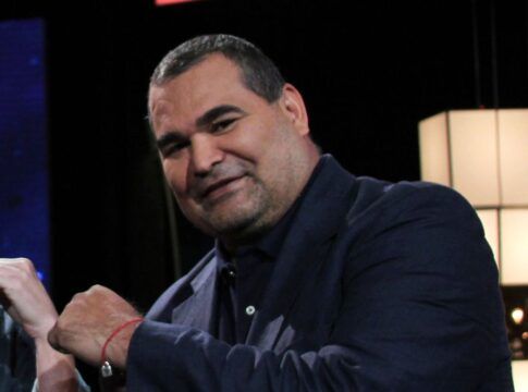 O ex-goleiro paraguaio José Luis Chilavert, um homem de pele clara e porte robusto, sorri levemente para a câmera. Ele veste um blazer azul-marinho sobre uma camisa social da mesma cor. No pulso direito, carrega uma pulseira de fio vermelho. À esquerda, parte do rosto de outro homem sorridente é visível, e o fundo escuro destaca luminárias cúbicas brancas à direita.