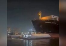 Registro noturno de um grande navio cargueiro escuro posicionado muito próximo a uma balsa de transporte de passageiros em um canal. A iluminação é artificial, vinda da embarcação e das luzes da cidade ao fundo, com a água refletindo o brilho das luzes.