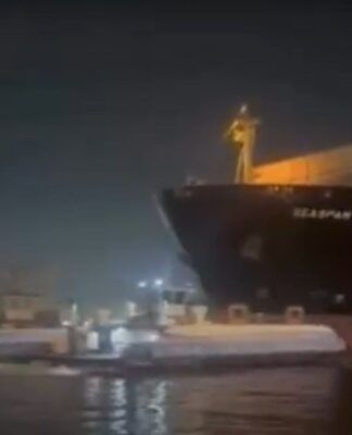 Registro noturno de um grande navio cargueiro escuro posicionado muito próximo a uma balsa de transporte de passageiros em um canal. A iluminação é artificial, vinda da embarcação e das luzes da cidade ao fundo, com a água refletindo o brilho das luzes.