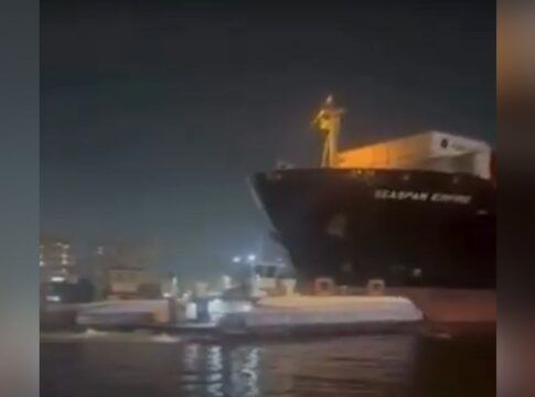 Registro noturno de um grande navio cargueiro escuro posicionado muito próximo a uma balsa de transporte de passageiros em um canal. A iluminação é artificial, vinda da embarcação e das luzes da cidade ao fundo, com a água refletindo o brilho das luzes.