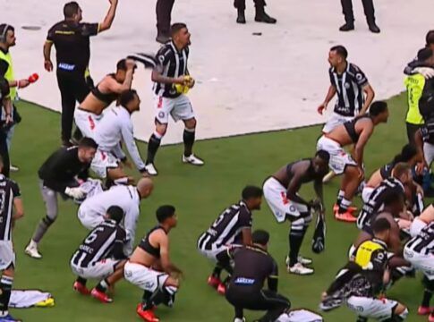 Jogadores e comissão técnica de um time de futebol, vestindo uniformes de listras verticais pretas e brancas com calções brancos, celebram intensamente em campo. Alguns atletas estão ajoelhados ou agachados no gramado, enquanto outros correm e agitam camisas no ar. Ao fundo, profissionais de imprensa com coletes e microfones acompanham a movimentação na borda do campo.