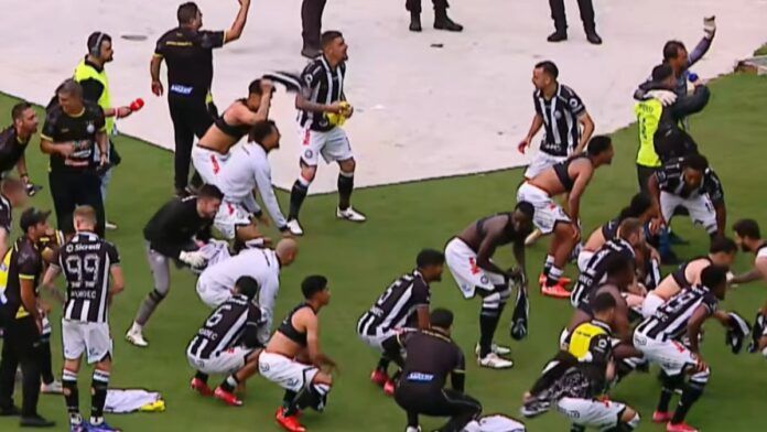 Jogadores e comissão técnica de um time de futebol, vestindo uniformes de listras verticais pretas e brancas com calções brancos, celebram intensamente em campo. Alguns atletas estão ajoelhados ou agachados no gramado, enquanto outros correm e agitam camisas no ar. Ao fundo, profissionais de imprensa com coletes e microfones acompanham a movimentação na borda do campo.