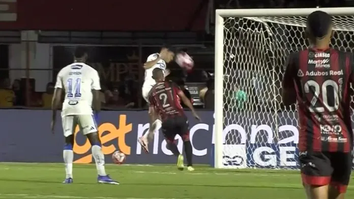 Registro de um lance de jogo de futebol à noite. Um jogador de uniforme branco salta para cabecear a bola em direção ao gol, enquanto um defensor de uniforme listrado em vermelho e preto tenta impedir a jogada. Em primeiro plano, as costas de outro jogador do mesmo time listrado, vestindo a camisa número 20, ocupam o lado direito da imagem. Ao fundo, vê-se a rede do gol e placas de publicidade no nível do gramado.