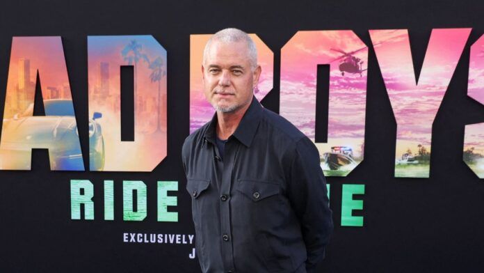 O ator Eric Dane, um homem de pele clara e cabelos curtos grisalhos, posa para uma foto em um tapete vermelho. Ele veste uma camisa social azul-marinho de botões. Ao fundo, há um painel preto com letras grandes coloridas que formam parte do título do filme 