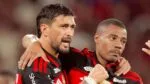 Dois jogadores de futebol do Flamengo estão em um momento de interação no campo. O jogador à esquerda, com barba e cabelo curto, está com o braço sobre o ombro do companheiro e parece falar algo com uma expressão intensa. O jogador à direita, com cabelo raspado, mantém uma expressão mais contida e séria. Ambos vestem a camisa rubro-negra (listras horizontais pretas e vermelhas) com o patrocínio 