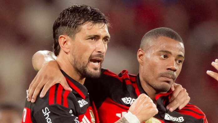 Dois jogadores de futebol do Flamengo estão em um momento de interação no campo. O jogador à esquerda, com barba e cabelo curto, está com o braço sobre o ombro do companheiro e parece falar algo com uma expressão intensa. O jogador à direita, com cabelo raspado, mantém uma expressão mais contida e séria. Ambos vestem a camisa rubro-negra (listras horizontais pretas e vermelhas) com o patrocínio 