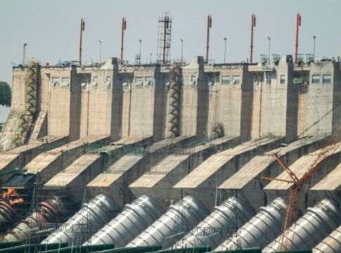 Vista externa da Usina Hidrelétrica de Belo Monte, mostrando a imensa estrutura de concreto da barragem. Na base, destacam-se os grandes condutos forçados cilíndricos e prateados que levam a água para as turbinas. No topo da estrutura, há diversas torres metálicas laranjas alinhadas, e ao fundo, à esquerda, uma área verde com árvores sob um céu claro.