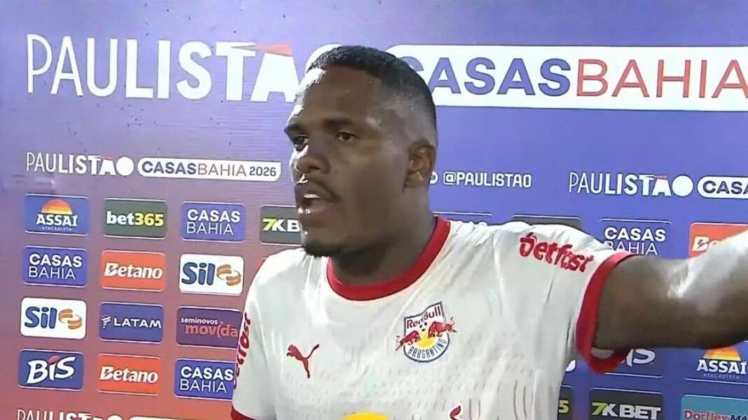 Um jogador de futebol negro, vestindo a camisa branca do Red Bull Bragantino, concede uma entrevista após uma partida. Ele está de perfil parcial, com a boca aberta como se estivesse falando e o braço esquerdo estendido. Ao fundo, há um painel de patrocinadores com as marcas 