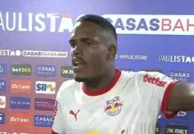 Um jogador de futebol negro, vestindo a camisa branca do Red Bull Bragantino, concede uma entrevista após uma partida. Ele está de perfil parcial, com a boca aberta como se estivesse falando e o braço esquerdo estendido. Ao fundo, há um painel de patrocinadores com as marcas 