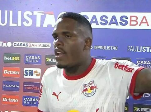 Um jogador de futebol negro, vestindo a camisa branca do Red Bull Bragantino, concede uma entrevista após uma partida. Ele está de perfil parcial, com a boca aberta como se estivesse falando e o braço esquerdo estendido. Ao fundo, há um painel de patrocinadores com as marcas 