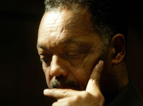 Close-up do rosto de um homem negro de cabelos grisalhos e bigode. Ele está com os olhos baixos, expressão séria e pensativa, com o dedo indicador encostado no queixo. O fundo está escuro e a iluminação foca em seu rosto.