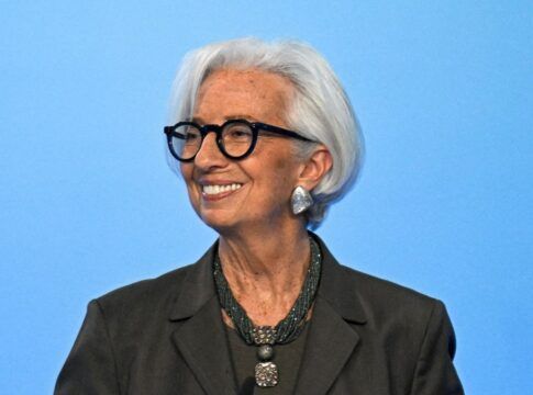 Uma mulher de cabelos brancos curtos e óculos de armação preta sorri para o lado. Ela veste um blazer escuro, um colar de várias voltas com um pingente quadrado e brincos prateados grandes. O fundo é de um azul claro uniforme.