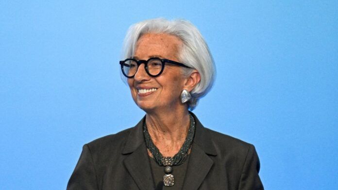 Uma mulher de cabelos brancos curtos e óculos de armação preta sorri para o lado. Ela veste um blazer escuro, um colar de várias voltas com um pingente quadrado e brincos prateados grandes. O fundo é de um azul claro uniforme.