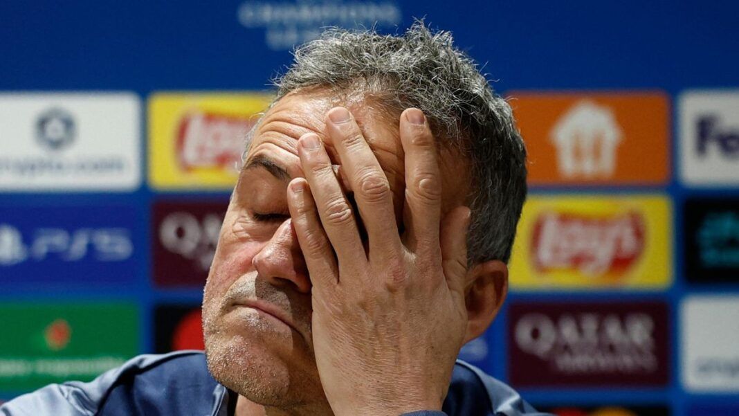 Um homem de pele clara e cabelos curtos grisalhos aparece em um plano fechado, com os olhos fechados e a mão cobrindo parte do rosto em um gesto de cansaço ou frustração. Ele veste uma peça de roupa azul. Ao fundo, há um painel desfocado com logotipos de patrocinadores e competições esportivas.