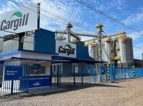 Vista externa de uma unidade industrial da Cargill sob um céu azul com nuvens esparsas. Em primeiro plano, há uma guarita branca e azul com uma placa indicando 