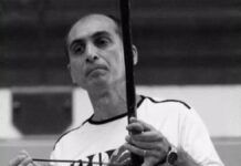 Fotografia em preto e branco de um homem de meia-idade, calvo no topo da cabeça, com expressão serena. Ele veste uma camiseta clara e segura um berimbau na posição vertical, com a mão esquerda no instrumento e a direita segurando a baqueta.