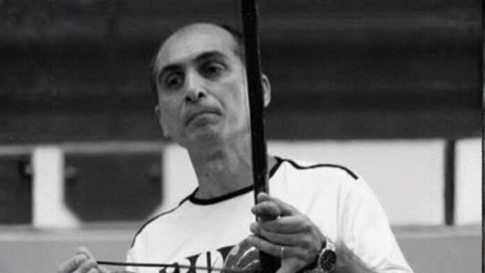 Fotografia em preto e branco de um homem de meia-idade, calvo no topo da cabeça, com expressão serena. Ele veste uma camiseta clara e segura um berimbau na posição vertical, com a mão esquerda no instrumento e a direita segurando a baqueta.