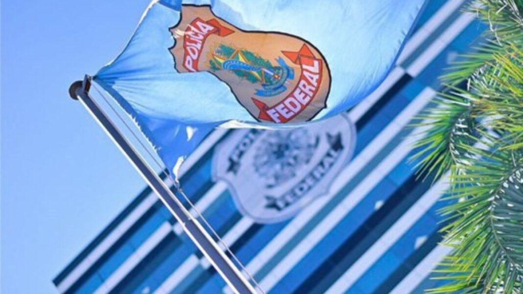 Bandeira azul clara da Polícia Federal hasteada e ondulando ao vento sob um céu azul. Ao fundo, de forma desfocada, vê-se a fachada de um prédio moderno com vidros espelhados e outro logotipo da instituição. À direita, parte das folhas de uma palmeira emoldura a cena.