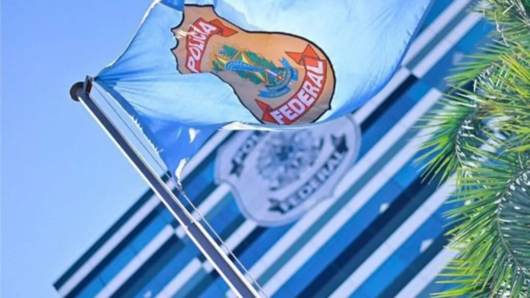 Bandeira azul clara da Polícia Federal hasteada e ondulando ao vento sob um céu azul. Ao fundo, de forma desfocada, vê-se a fachada de um prédio moderno com vidros espelhados e outro logotipo da instituição. À direita, parte das folhas de uma palmeira emoldura a cena.