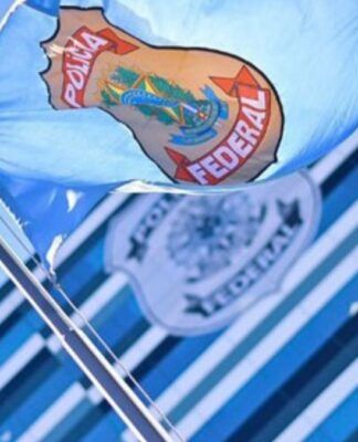 Bandeira azul clara da Polícia Federal hasteada e ondulando ao vento sob um céu azul. Ao fundo, de forma desfocada, vê-se a fachada de um prédio moderno com vidros espelhados e outro logotipo da instituição. À direita, parte das folhas de uma palmeira emoldura a cena.