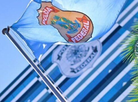 Bandeira azul clara da Polícia Federal hasteada e ondulando ao vento sob um céu azul. Ao fundo, de forma desfocada, vê-se a fachada de um prédio moderno com vidros espelhados e outro logotipo da instituição. À direita, parte das folhas de uma palmeira emoldura a cena.