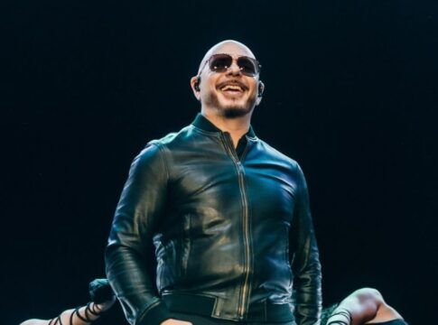 O cantor Pitbull, um homem calvo de pele clara, sorri enquanto se apresenta em um palco. Ele usa óculos de sol escuros, uma jaqueta de couro preta fechada e fones de ouvido de monitoramento (in-ear). O fundo é escuro, com luzes de palco focadas nele, e parte dos braços de dançarinas é visível na parte inferior da imagem.