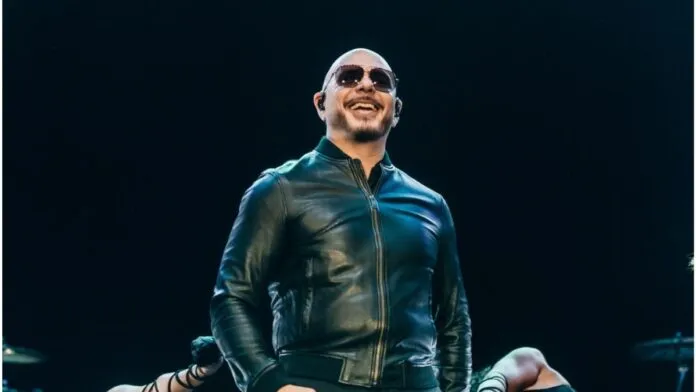 O cantor Pitbull, um homem calvo de pele clara, sorri enquanto se apresenta em um palco. Ele usa óculos de sol escuros, uma jaqueta de couro preta fechada e fones de ouvido de monitoramento (in-ear). O fundo é escuro, com luzes de palco focadas nele, e parte dos braços de dançarinas é visível na parte inferior da imagem.