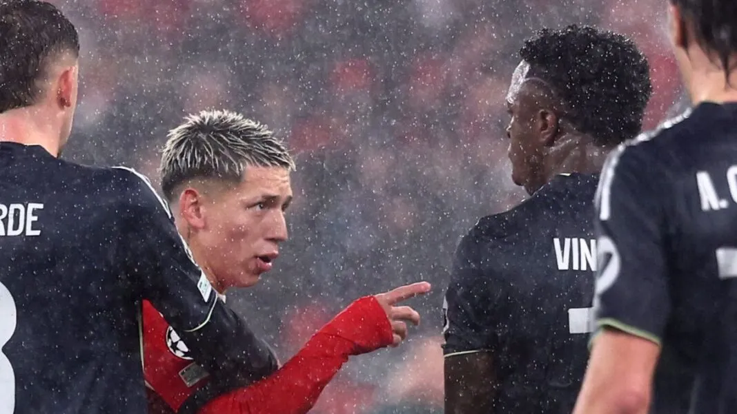 Sob chuva intensa, dois jogadores de futebol interagem no gramado durante uma partida noturna. O jogador à esquerda, com cabelo loiro curto e descolorido, veste um uniforme vermelho de mangas longas e aponta o dedo indicador em direção ao outro atleta. O jogador à direita, de costas para a câmera, veste um uniforme preto com o nome 