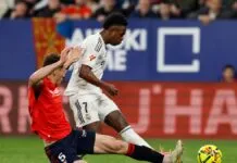 O jogador Vinícius Júnior, vestindo o uniforme branco do Real Madrid, está em movimento com a bola durante uma partida de futebol. Ele é confrontado por um adversário de uniforme vermelho e calção azul, que tenta desarmá-lo com um carrinho. Ao fundo, é possível ver a arquibancada com torcedores e placas de publicidade coloridas.