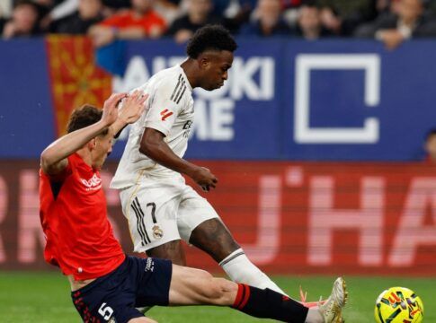 O jogador Vinícius Júnior, vestindo o uniforme branco do Real Madrid, está em movimento com a bola durante uma partida de futebol. Ele é confrontado por um adversário de uniforme vermelho e calção azul, que tenta desarmá-lo com um carrinho. Ao fundo, é possível ver a arquibancada com torcedores e placas de publicidade coloridas.