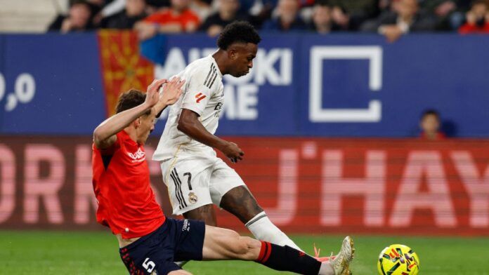 O jogador Vinícius Júnior, vestindo o uniforme branco do Real Madrid, está em movimento com a bola durante uma partida de futebol. Ele é confrontado por um adversário de uniforme vermelho e calção azul, que tenta desarmá-lo com um carrinho. Ao fundo, é possível ver a arquibancada com torcedores e placas de publicidade coloridas.